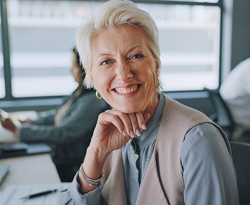 Das Bild zeigt eine strahlende, ältere Frau im Büro, die mit einem warmen Lächeln und professioneller Ausstrahlung die Betrachter anspricht.  Im Detail ist eine reife Frau mit kurzem, weißem Haar der Mittelpunkt des Bildes; sie trägt eine hellblaue Bluse mit einer beigen Weste, die ihrem Outfit eine elegante Note verleiht. Ihr Lächeln wirkt herzlich und einladend, während sie ihre Hand unter ihr Kinn legt, was eine nachdenkliche, aber selbstbewusste Haltung unterstreicht. Im Hintergrund sind Büroarbeitsplätze mit Monitoren und anderen Personen zu erkennen, was auf eine belebte, aber professionelle Arbeitsumgebung hindeutet. Die allgemeine Atmosphäre ist positiv und professionell, vermittelt ein Gefühl von Kompetenz und Erfahrung. Auf dem Tisch vor ihr liegen Dokumente und ein Stift, was auf ihre Beteiligung am Arbeitsgeschehen hinweist. Das Bild strahlt eine Stimmung von Vertrauen und Zuversicht aus.