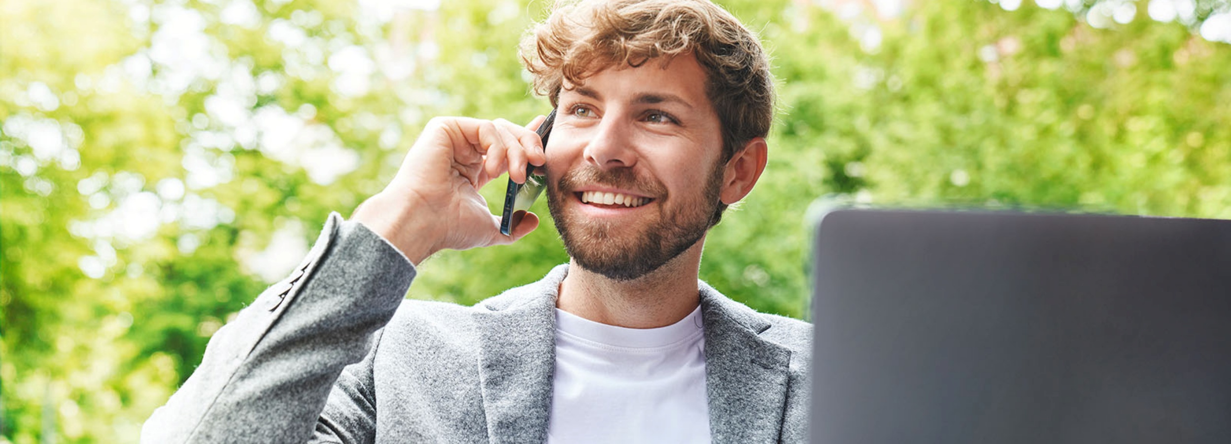 Ein gutaussehender Mann telefoniert lächelnd im Freien vor einem Laptop und einer grünen Baumkulisse.  Das Bild zeigt einen jungen Mann mit lockigem, hellbraunem Haar und einem gepflegten Bart, der ein entspanntes Lächeln zeigt, während er ein Smartphone an sein Ohr hält. Er trägt ein helles T-Shirt unter einem grauen Sakko mit Knöpfen an den Ärmeln, was auf einen smart-casual Stil hindeutet. Vor ihm befindet sich ein Laptop, dessen Bildschirm unsichtbar ist. Der Hintergrund ist ein verschwommener, grüner Bereich, der wahrscheinlich von Bäumen stammt, was auf eine natürliche, vielleicht parkähnliche Umgebung hindeutet. Die Atmosphäre wirkt hell und positiv, unterstrichen durch das Lächeln des Mannes und die sonnige Farbgebung des Hintergrunds.