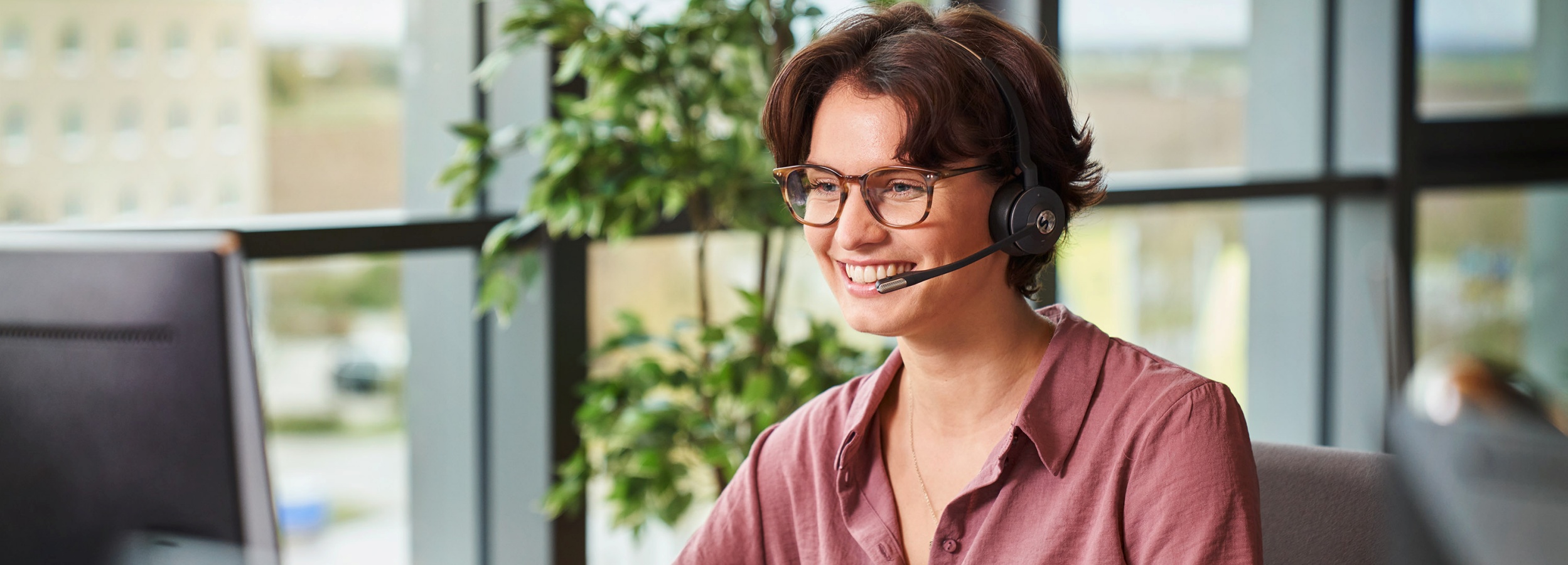Eine lächelnde Frau mit Headset sitzt in einem Büro und arbeitet an einem Computer.  Das Bild zeigt eine Frau mit braunen, gewellten Haaren und Brille, die ein Headset trägt und vor einem Computer sitzt. Sie trägt ein rosafarbenes Hemd und lächelt, was eine positive und freundliche Atmosphäre vermittelt. Im Hintergrund ist ein großes Fenster zu sehen, das einen Blick auf ein Gebäude und etwas Grün ermöglicht. Eine Pflanze steht in der Nähe des Fensters und trägt zur lebendigen und hellen Stimmung des Raumes bei. Ein Teil des Computerbildschirms ist ebenfalls im Bild sichtbar. Die allgemeine Stimmung des Bildes ist professionell und einladend, was darauf hindeutet, dass die Frau entweder in einem Kundenservice oder in einer ähnlichen Rolle arbeitet, die Kommunikation erfordert.