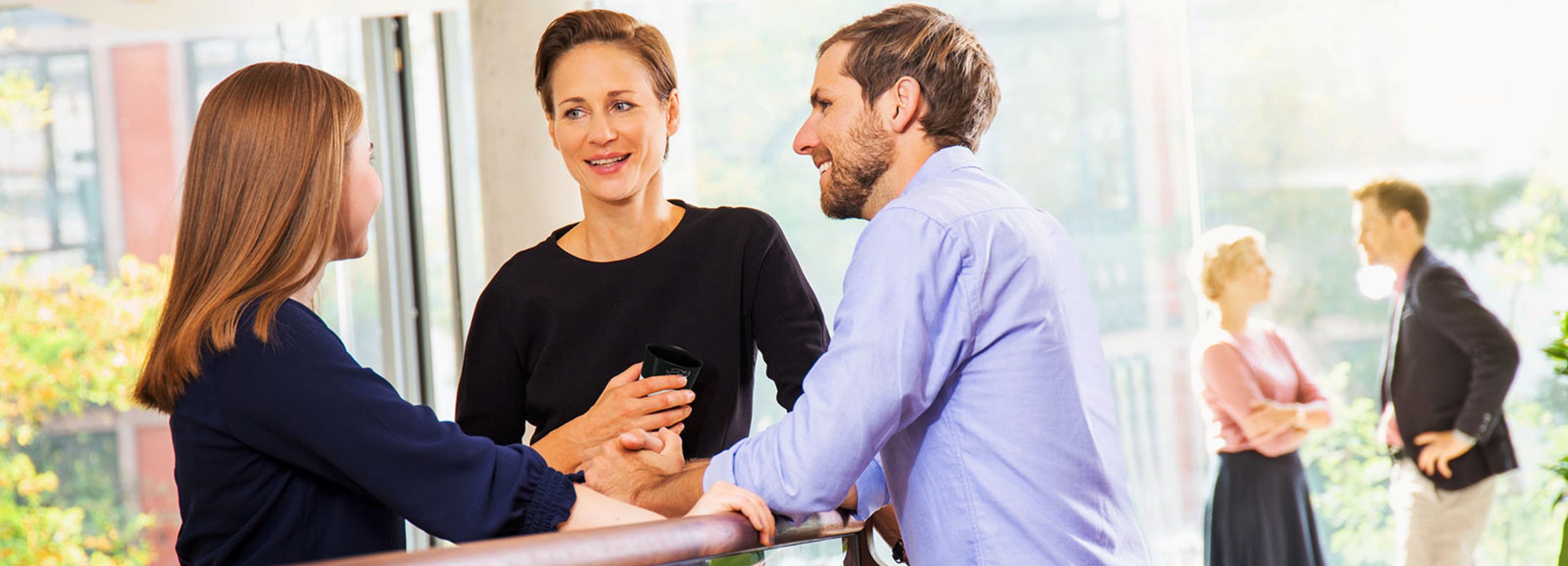 Das Bild zeigt eine lebhafte Unterhaltung zwischen Kollegen in einem hellen, modernen Bürogebäude.  Im Vordergrund stehen drei Personen, die sich angeregt unterhalten. Eine junge Frau mit langen, glatten, hellbraunen Haaren und einem dunkelblauen Oberteil steht links und scheint aufmerksam zuzuhören. In der Mitte steht eine Frau mit kurzen, hellbraunen Haaren, die ein schwarzes Oberteil trägt und einen schwarzen Becher in der Hand hält. Rechts lehnt sich ein Mann mit Bart und hellblauem Hemd an ein Geländer und beteiligt sich aktiv an der Konversation. Alle drei Personen wirken entspannt und lächeln, was eine positive und freundliche Atmosphäre vermittelt.  Im Hintergrund sind verschwommen zwei weitere Personen zu sehen, die ebenfalls in ein Gespräch vertieft sind. Eine Frau mit blonden Haaren trägt ein rosa Oberteil und einen dunklen Rock, während der Mann einen dunklen Anzug trägt. Der helle Hintergrund mit Fenstern und natürlichem Licht trägt zur offenen und luftigen Stimmung des Bildes bei. Das gesamte Bild vermittelt den Eindruck eines dynamischen und kooperativen Arbeitsumfelds.