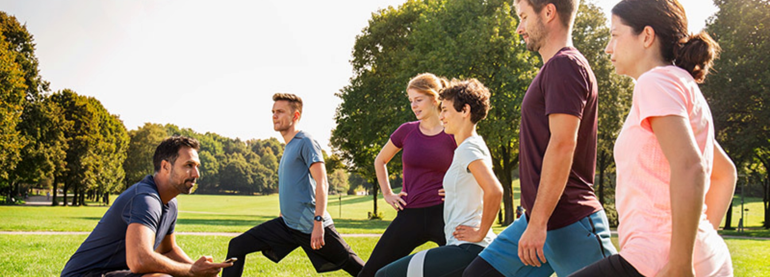 Das Bild zeigt eine Gruppe von Menschen, die im Freien unter Anleitung eines Trainers Dehnübungen machen.  Im Detail zeigt das Bild eine Gruppe von fünf Personen, die sich in einem Park befinden. Vier von ihnen stehen in einer Reihe und führen Dehnübungen aus, während der fünfte, offensichtlich der Trainer, daneben sitzt und ein Smartphone in der Hand hält, während er ihnen Anweisungen gibt. Die Personen tragen Sportkleidung unterschiedlicher Farben, was eine gewisse Vielfalt in der Gruppe andeutet. Der Trainer, der sich in der Nähe der Gruppe befindet, scheint die Übungen zu überwachen oder zu leiten, möglicherweise mithilfe der Informationen auf seinem Smartphone. Der Hintergrund besteht aus einer grünen Rasenfläche und Bäumen, was eine natürliche und entspannte Atmosphäre schafft. Das Wetter scheint sonnig zu sein, was die Stimmung des Bildes positiv und aktiv macht. Die Szene vermittelt ein Gefühl von Gesundheit, Aktivität und Gemeinschaft, da die Gruppe zusammen trainiert.