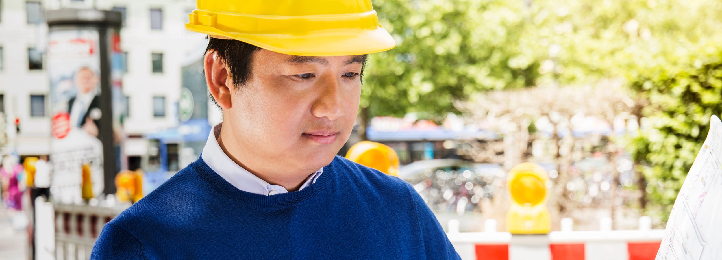 Das Bild zeigt einen Bauleiter, der aufmerksam einen Bauplan studiert.  Im Detail zeigt das Bild einen asiatischen Mann mit dunklen Haaren und hellbrauner Haut, der einen gelben Bauhelm trägt. Er trägt einen blauen Pullover über einem weißen Hemd. Der Mann blickt konzentriert auf einen Bauplan, den er in seinen Händen hält. Im Hintergrund ist eine städtische Szene zu erkennen, mit Bäumen, Gebäuden und einem Baustellenzaun mit gelben Warnbaken, die auf eine Baustelle oder eine Straßensperrung hindeuten. Die Atmosphäre wirkt geschäftig und professionell, da der Bauleiter sich auf seine Aufgaben konzentriert. Das Bild vermittelt den Eindruck von Planung und Präzision im Bauwesen.