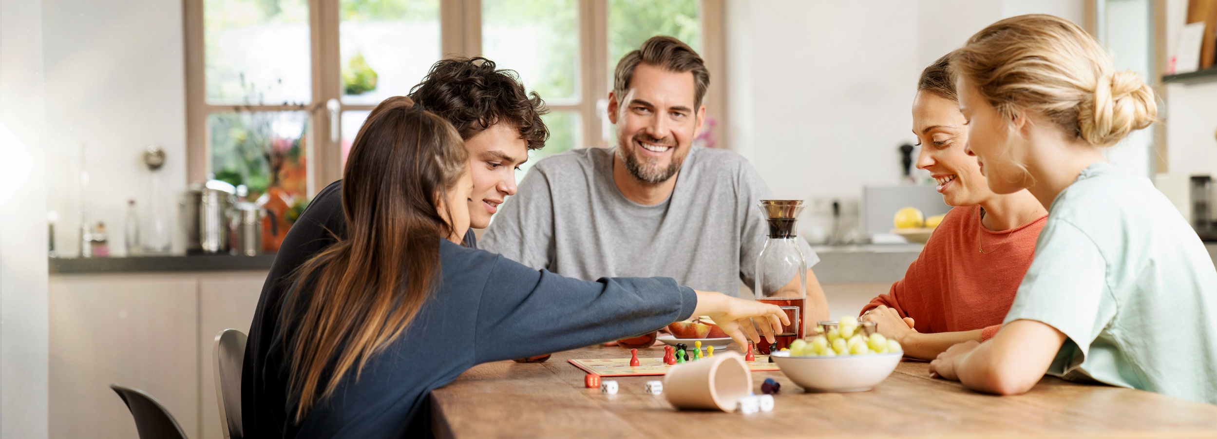 Eine glückliche Familie sitzt an einem Holztisch in einer Küche und spielt ein Brettspiel.  Auf dem Bild sind fünf Personen zu sehen, die an einem Tisch sitzen. Eine junge Frau mit langen, braunen Haaren und einem dunkelblauen Pullover ist im Begriff eine Spielfigur zu bewegen. Neben ihr sitzt ein junger Mann mit lockigen, braunen Haaren und einem grauen T-Shirt. Hinter den beiden sitzt ein Mann mit Bart, ebenfalls in einem grauen T-Shirt, der lächelt in die Kamera. Rechts davon sitzt eine Frau mit braunen Haaren und einem orangefarbenen Pullover, die ebenfalls lächelt. Ganz rechts sitzt ein junges Mädchen mit blonden Haaren, die zu einem Dutt gebunden sind, und einem hellgrünen T-Shirt.  Auf dem Tisch befindet sich ein Brettspiel mit bunten Spielfiguren, Würfeln und einem Würfelbecher. Außerdem stehen dort eine Karaffe mit Getränk, ein Glas, eine Schale mit Weintrauben und ein Teller mit Äpfeln.  Die Küche im Hintergrund ist hell und modern eingerichtet, mit weißen Schränken und Edelstahlgeräten. Die Atmosphäre des Bildes ist warm und einladend, und vermittelt das Gefühl von Familienglück und guter Laune.