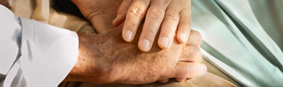 Das Bild zeigt zwei Hände, die sich halten, wobei die jüngere Hand die ältere Hand stützt und eine Stimmung von Trost und Unterstützung vermittelt.  Im Detail zeigt das Bild eine Nahaufnahme zweier Hände. Die darüber liegende Hand scheint jünger zu sein und hat eine glattere Haut. Sie bedeckt und hält die Hand darunter. Die darunter liegende Hand ist älter, mit sichtbaren Falten und Altersflecken, was auf ein höheres Alter hindeutet. Man sieht einen Teil eines weißen Hemdes oder Kittels auf der linken Seite, was darauf hindeuten könnte, dass die Person in einem medizinischen Beruf arbeitet. Die Hände liegen auf einer Textur, die an ein beigefarbenes Tuch oder eine Decke erinnert. Im Hintergrund ist ein hellbläuliches, glänzendes Material zu sehen, möglicherweise ein Krankenbettlaken. Die Beleuchtung ist weich und warm, was zu einer intimen und ruhigen Atmosphäre beiträgt. Die Stimmung ist ruhig und könnte Gefühle von Mitgefühl, Sorge oder tröstender Unterstützung vermitteln.