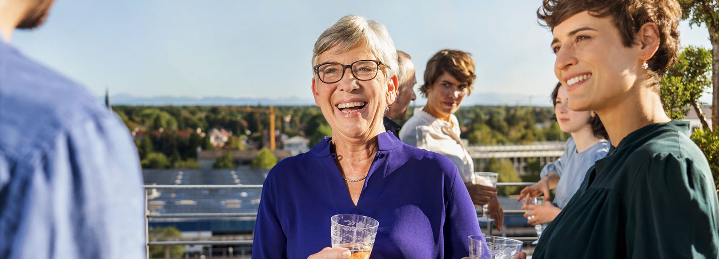Das Bild zeigt eine Gruppe von Menschen, die sich auf einer Dachterrasse unterhalten und dabei eine ausgelassene und freundliche Atmosphäre genießen.  Im Detail zeigt das Bild eine Gruppe von etwa sechs Personen, die auf einer Dachterrasse stehen. Im Vordergrund ist eine ältere Frau mit kurzen, grauen Haaren und Brille zu sehen, die herzlich in die Kamera lacht und ein Glas in der Hand hält. Sie trägt eine blaue Bluse. Rechts von ihr steht eine jüngere Frau mit dunklen, lockigen Haaren, die ebenfalls lächelt und ein grünes Oberteil trägt. Im Hintergrund sind weitere Personen erkennbar, darunter ein Mann mit grauem Haar und eine junge Frau mit braunen Haaren, die in einem hellen Oberteil steht und ein Glas hält. Eine weitere junge Frau ist im Hintergrund ebenfalls zu sehen, etwas verdeckt. Links im Bild ist der Rücken einer weiteren Person in einem blauen Hemd zu sehen, die scheinbar ebenfalls Teil der Unterhaltung ist.  Die Dachterrasse bietet einen weiten Blick über eine Stadtlandschaft mit Bäumen und Gebäuden. Der Himmel ist hellblau und sonnig, was zur positiven und entspannten Stimmung des Bildes beiträgt. Die allgemeine Atmosphäre ist fröhlich und einladend, was darauf schließen lässt, dass die Personen eine angenehme Zeit miteinander verbringen.
