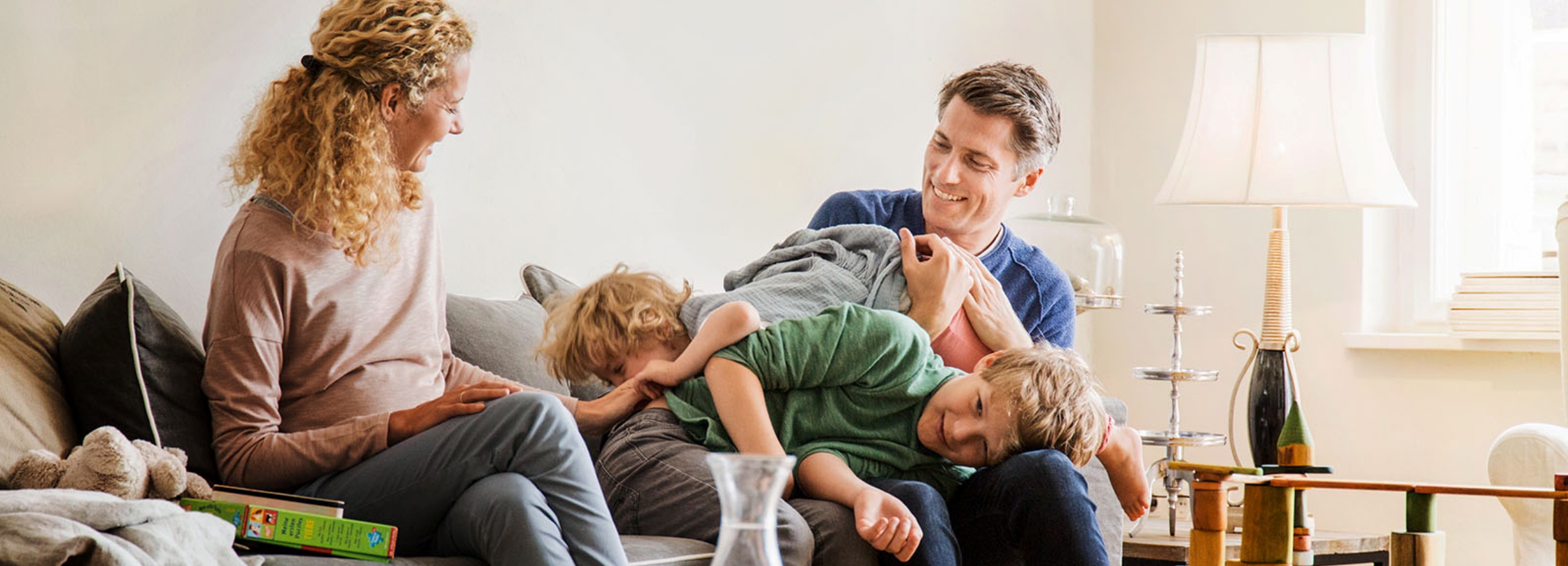 Das Bild zeigt eine Szene häuslicher Glück mit einer Familie, die sich auf einem Sofa entspannt und spielt.  Im Detail zeigt das Foto einen hellen und freundlichen Wohnraum. Auf dem Sofa sitzen zwei Erwachsene und zwei Kinder. Die Frau, mit lockigem blondem Haar, sitzt links und schaut lächelnd auf einen Laptop. Sie trägt ein hellrosa Langarmshirt und Jeans. Neben ihr auf dem Sofa liegen ein Kuscheltier und einige Kinderbücher. Der Mann, mit kurzen braunen Haaren, sitzt rechts und ist aktiv mit den beiden Jungen beschäftigt, die auf ihm herumtollen. Er trägt ein blaues Langarmshirt. Einer der Jungen hat blonde Haare und liegt auf dem Rücken des Vaters, während der andere Junge, ebenfalls blond, auf dem Schoß des Vaters liegt und ihn umarmt.  Im Hintergrund ist eine weiße Wand sichtbar, vor der ein Beistelltisch mit einer Lampe, einer Etagere und einigen Dekorationsgegenständen steht. Ein Fenster sorgt für natürliches Licht im Raum. Die Atmosphäre ist warm, liebevoll und verspielt, was durch die fröhlichen Gesichtsausdrücke und die lebhafte Interaktion zwischen den Familienmitgliedern unterstrichen wird. Das Bild vermittelt ein Gefühl von Geborgenheit und familiärer Verbundenheit.