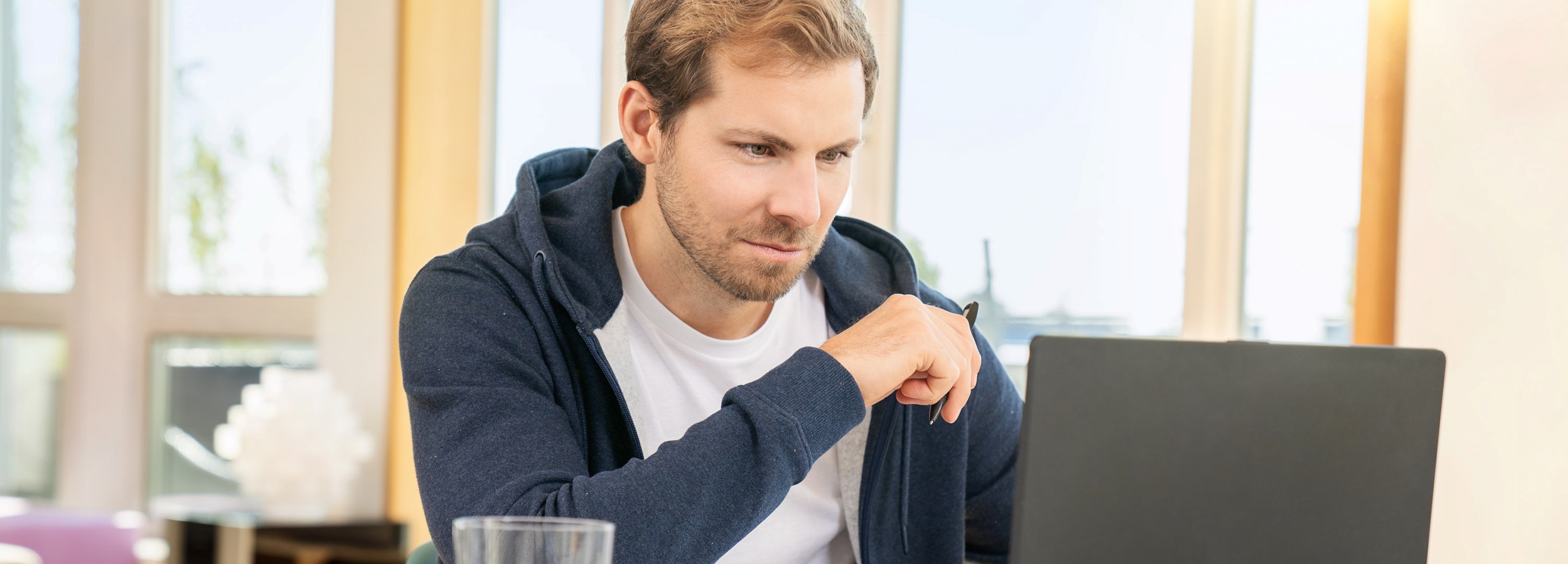 Das Bild zeigt einen Mann, der an seinem Laptop arbeitet, vermutlich von zu Hause aus.  Detailliert betrachtet, ist ein Mann mittleren Alters zu sehen, der vor einem Laptop sitzt. Er trägt ein weißes T-Shirt unter einem dunkelblauen Kapuzenpullover. Seine Haltung und sein Blick deuten auf Konzentration und Beschäftigung mit dem Bildschirm hin. In seiner Hand hält er einen Stift.  Im Hintergrund befindet sich ein Fenster, das viel Tageslicht hereinlässt, was eine helle und freundliche Atmosphäre schafft. Außerhalb des Fensters sind verschwommen Bäume und Gebäude erkennbar. Die Szene wirkt ruhig und produktiv, was die Stimmung eines typischen Homeoffice-Szenarios vermittelt. Ein Glas steht ebenfalls im Vordergrund, was auf eine entspannte Arbeitsumgebung hindeutet.