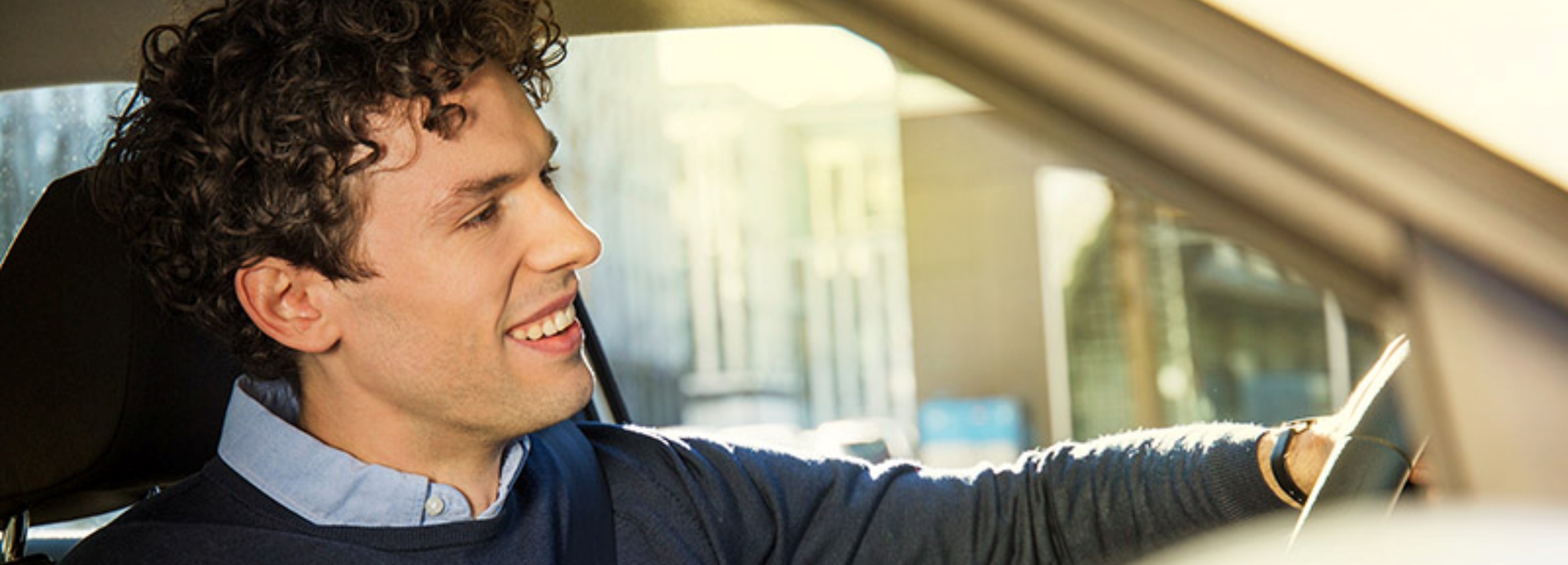 Ein junger Mann mit lockigem Haar sitzt am Steuer eines Autos und blickt mit einem Lächeln auf dem Gesicht nach außen.  Das Bild zeigt das Profil eines jungen Mannes mit lockigem, dunkelbraunem Haar. Er trägt ein hellblaues Hemd unter einem dunkelblauen Pullover und einen Sicherheitsgurt. Er sitzt am Steuer eines Autos und hält das Lenkrad fest. Der Mann blickt nach links aus dem Fenster und lächelt dabei, was auf eine positive oder interessierte Stimmung hindeutet. Das Sonnenlicht scheint hell auf sein Gesicht, während im Hintergrund Gebäude und möglicherweise Bäume zu sehen sind, die jedoch unscharf sind. Die Atmosphäre wirkt hell und freundlich, und die Stimmung ist optimistisch und entspannt.