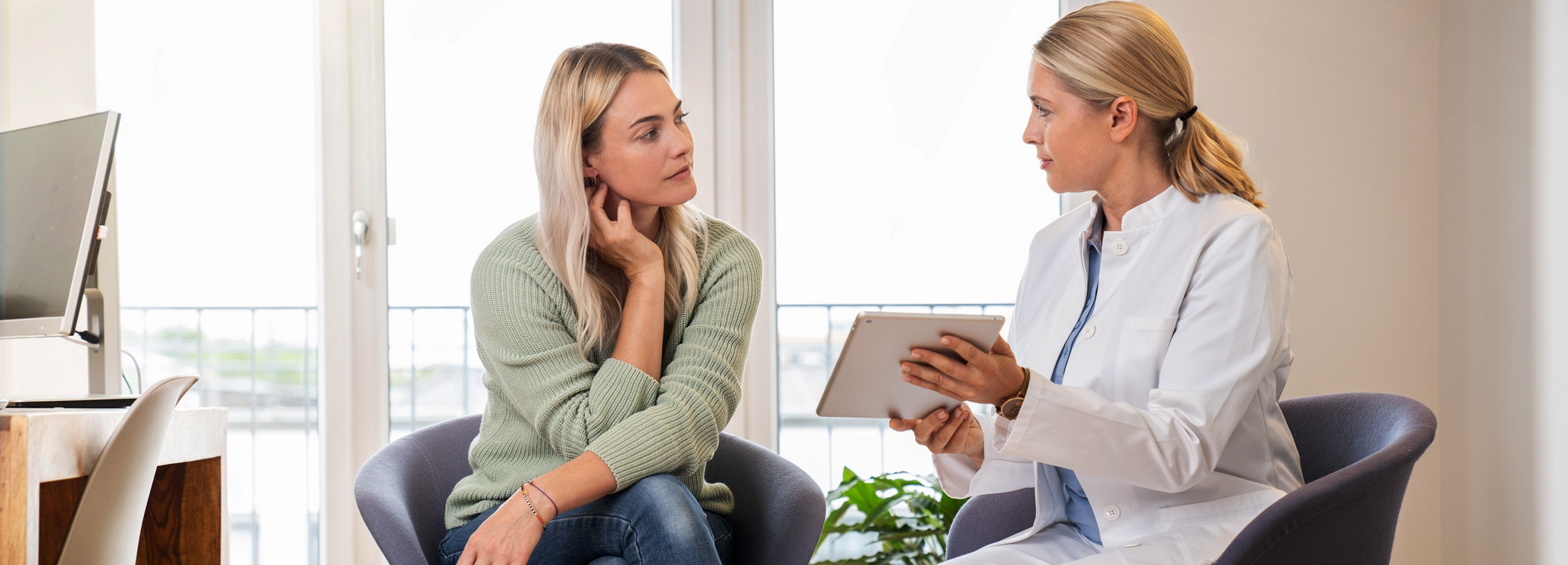 Eine Patientin und eine Ärztin sitzen in einem hellen Raum, die Ärztin hält ein Tablet und scheint etwas zu erklären.  Das Bild zeigt zwei Frauen in einem hellen, modern eingerichteten Raum. Die linke Frau, vermutlich die Patientin, sitzt in einem grauen Sessel und trägt einen hellgrünen Strickpullover und Jeans. Sie hat blondes, schulterlanges Haar und blickt nachdenklich zur anderen Frau. Ihre Hand liegt an ihrer Wange, was einen Anflug von Besorgnis oder Nachdenklichkeit suggeriert. Auf ihrem Handgelenk trägt sie mehrere Armbänder.  Die rechte Frau, vermutlich die Ärztin, trägt einen weißen Arztkittel über einem blauen Hemd. Sie hat ebenfalls blondes Haar, das zu einem Pferdeschwanz zusammengebunden ist. In ihren Händen hält sie ein Tablet und scheint der Patientin etwas zu erklären. Ihr Blick ist zugewandt und aufmerksam.  Zwischen den Frauen befindet sich eine ruhige und professionelle Atmosphäre, es könnte sich um eine Beratungs- oder Untersuchungssituation handeln. Im Hintergrund sind ein Fenster mit hellem Tageslicht, ein Schreibtisch mit einem Computer und ein weiterer Stuhl zu sehen, was den Eindruck eines Arztzimmers oder Beratungsraums verstärkt. Das Bild wirkt insgesamt beruhigend und professionell.