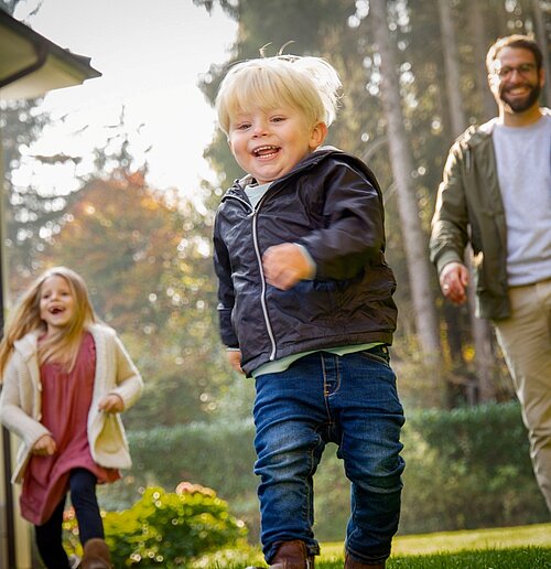 Das Bild zeigt eine glückliche Familie, die an einem sonnigen Tag draußen spielt.  Im Detail zeigt das Bild einen fröhlichen kleinen Jungen mit blonden Haaren, der in die Kamera rennt und dabei ausgelassen lacht. Er trägt eine blaue Jacke und Jeans. Hinter ihm und leicht links ist ein Mädchen mit langen braunen Haaren zu sehen, das ebenfalls lachend rennt; sie trägt ein rosafarbenes Kleid und eine helle Strickjacke. Im Hintergrund, leicht unscharf, steht ein Mann mit Bart, der auch lächelt; er trägt eine grüne Jacke und helle Hosen. Die Szene spielt sich im Freien ab, vermutlich in einem Garten oder Park, mit Bäumen und etwas Grün im Hintergrund. Die allgemeine Atmosphäre ist fröhlich und unbeschwert, die Stimmung ist positiv und voller Freude. Das Sonnenlicht trägt zur warmen und einladenden Atmosphäre bei.