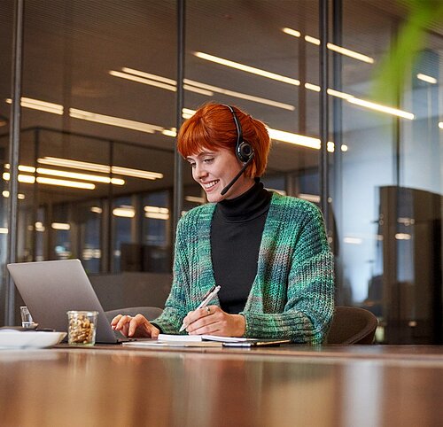 Gerne, hier ist die Bildbeschreibung:  Das Bild zeigt eine lächelnde Frau mit roten Haaren und einem Headset, die an einem Tisch sitzt und gleichzeitig an einem Laptop arbeitet und in ein Notizbuch schreibt.  Im Detail ist Folgendes zu sehen:  Person: Eine junge Frau mit auffällig roten Haaren, die zu einem lockeren Dutt hochgesteckt sind. Sie trägt ein Headset, was darauf hindeutet, dass sie möglicherweise in einem Kundenservice oder in einem ähnlichen Beruf arbeitet. Sie lächelt freundlich. Ihre Kleidung besteht aus einem schwarzen Rollkragenpullover und einer grün-blau gemusterten Strickjacke. Objekte: Auf dem Tisch vor ihr befinden sich ein Laptop, ein Notizbuch, ein Stift, ein Glas mit Inhalt (vermutlich Nüsse oder Snacks), ein Smartphone und einige Papiere. Der Tisch selbst ist aus poliertem Holz. Hintergrund: Der Hintergrund ist unscharf, aber man erkennt ein Bürogebäude mit Glaswänden und Leuchtstoffröhren. Atmosphäre und Stimmung: Die Atmosphäre wirkt professionell und freundlich. Die Frau scheint ihre Arbeit zu genießen. Das Bild strahlt eine positive und produktive Stimmung aus.  Insgesamt vermittelt das Bild den Eindruck einer engagierten und freundlichen Person, die in einem modernen Büro arbeitet.