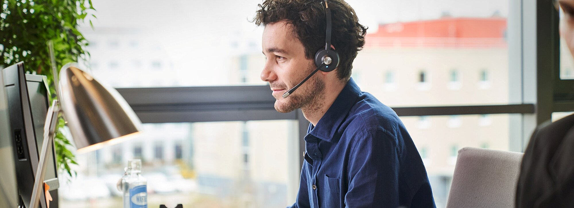 Das Bild zeigt einen Mann in einem Büro, der ein Headset trägt und vor einem Computer sitzt.  Im Detail zeigt das Bild einen Mann mit dunklem, lockigem Haar und leichtem Bart, der ein schwarzes Headset trägt. Er sitzt an einem Schreibtisch in einem Büro und blickt auf einen Computermonitor, der nur teilweise sichtbar ist. Er trägt ein blaues Hemd. Auf dem Schreibtisch befindet sich eine silberne Lampe, eine Flasche Wasser und ein Topf mit einer grünen Pflanze. Im Hintergrund ist ein Fenster mit Blick auf ein Stadtbild zu sehen. Ein Teil einer anderen Person ist am rechten Rand des Bildes erkennbar. Die Atmosphäre wirkt geschäftsmäßig und konzentriert. Die Stimmung ist ruhig und professionell.