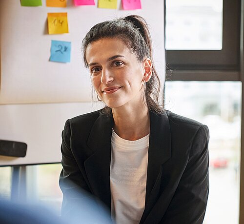 Das Bild zeigt eine lächelnde Geschäftsfrau in einem Büro-Setting.  Im Vordergrund ist eine Frau mit dunklen Haaren zu sehen, die zu einem Pferdeschwanz gebunden sind. Sie trägt Ohrringe, ein weißes T-Shirt mit einem kaum erkennbaren Aufdruck und darüber einen schwarzen Blazer. Die Frau lächelt leicht und blickt dem Betrachter direkt entgegen. Im Hintergrund ist eine weiße Tafel mit bunten Haftnotizen erkennbar, was auf eine Büro- oder Besprechungssituation hindeutet. Ein Fenster mit Blick auf eine unscharfe städtische Szene mit einem roten Auto ist ebenfalls sichtbar. Die Atmosphäre des Bildes ist professionell und positiv, vermittelt aber gleichzeitig eine gewisse Zugänglichkeit durch das Lächeln der Frau. Der Fokus liegt klar auf der Frau, während der Hintergrund eher unscharf gehalten ist, um die Aufmerksamkeit auf sie zu lenken.